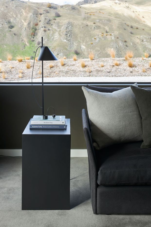 A refined living room vignette featuring a dark upholstered lounge chair with grey cushions, a slim black side table topped with books and a small object, and a minimalist black floor lamp. A large horizontal window frames a sparse, mountainous landscape, while grey carpet and a dark lower wall create a muted, contemporary atmosphere.