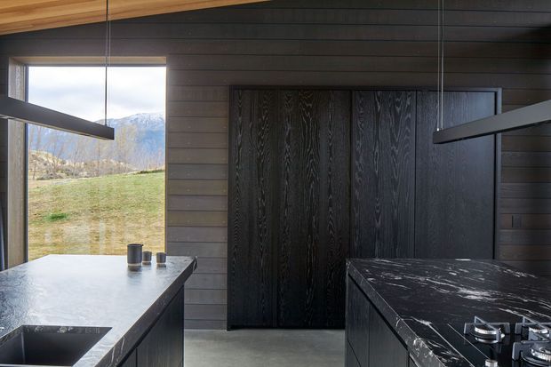 A contemporary kitchen with dark horizontal timber wall panelling, matching dark cabinetry, and dramatic black veined stone benchtops. Linear pendant lights hang above the island and cooking area. Floor-to-ceiling glazing opens to a grassy rural landscape and mountains, while warm timber ceiling detailing adds contrast.