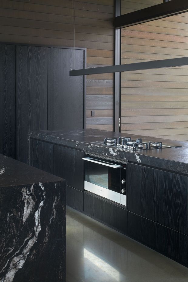 A dark, sophisticated contemporary kitchen featuring black vertically grained cabinetry, a dramatic dark stone island and benchtop with prominent white veining, integrated oven, gas cooktop, timber wall panelling, and black-framed glazing. A linear suspended pendant light spans the cooking area.