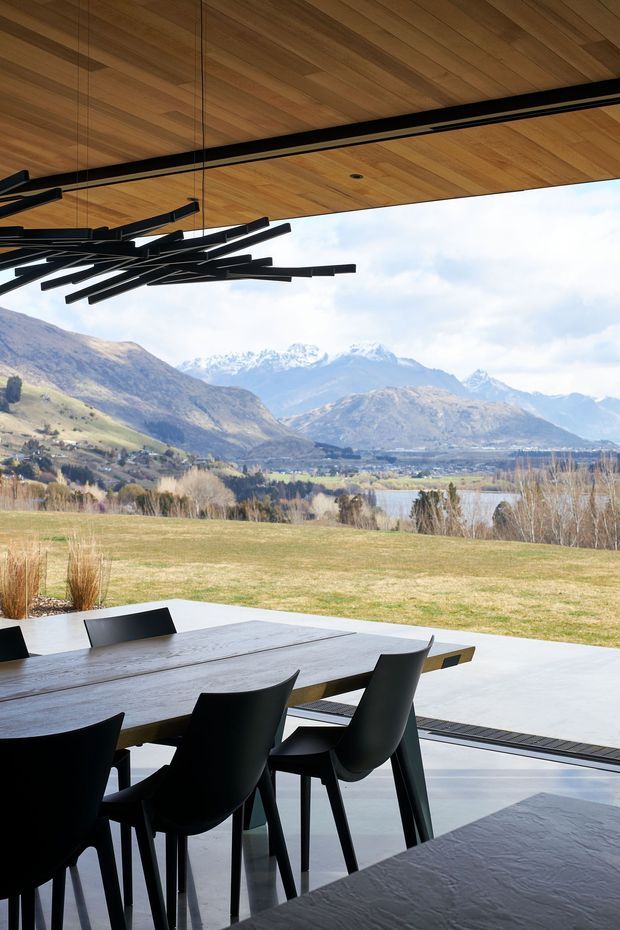 A contemporary dining area beside expansive floor-to-ceiling glazing, opening onto a terrace and a broad alpine mountain and lake landscape. The interior features a long dark timber dining table, black sculptural dining chairs, a warm timber-lined ceiling, and a striking linear black pendant light. The setting is airy and refined, with strong indoor-outdoor connection and panoramic views.