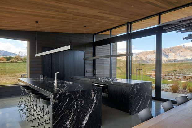 A contemporary kitchen with two large black marble islands, dark cabinetry, integrated sinks and taps, and bar seating. Floor-to-ceiling black-framed glazing provides expansive views of a grassy rural landscape and mountains. The room features a warm timber-lined ceiling, dark walls, linear pendant lighting, and polished concrete flooring.