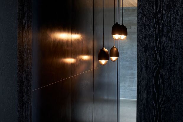 A moody architectural interior detail showing three suspended pendant lights with warm glowing bulbs against a dark, vertically panelled wall. A rough grey concrete wall and polished grey floor are visible through the opening, creating a dramatic, minimalist-industrial atmosphere.