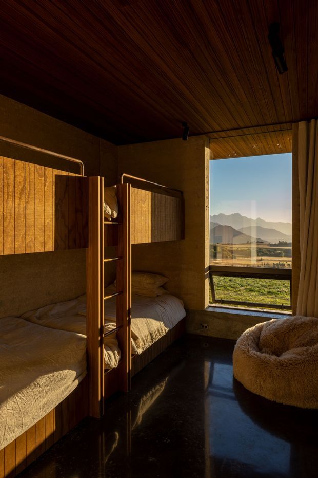 A compact, warmly lit bedroom with two built-in bunk beds in rich timber joinery, textured neutral bedding, a dark polished floor, and a large window overlooking an open rural landscape and mountains. The room has a timber-lined ceiling, minimal lighting, and a plush boucle lounge chair, creating a cozy cabin-like atmosphere.