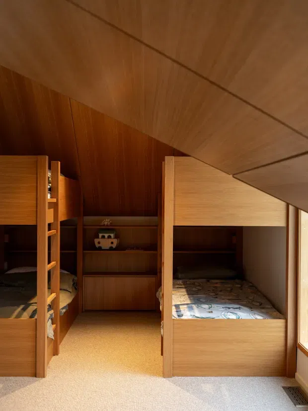 Compact children's bedroom with built-in wooden bunk beds, integrated shelving and cabinetry, warm wood wall and ceiling panelling, and a light carpeted floor. Strong natural light enters through the window on the right, creating a warm, cozy timber-lined interior.