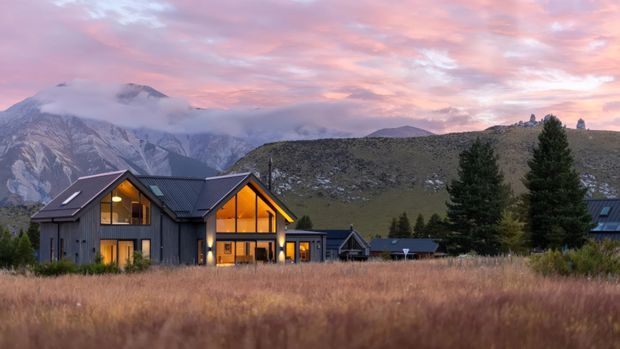 A modern mountain residence with dark vertical cladding, steep pitched roofs, and warm amber-lit glazing, set among tall grasses and evergreen trees with dramatic misty alpine mountains rising behind it at sunset.