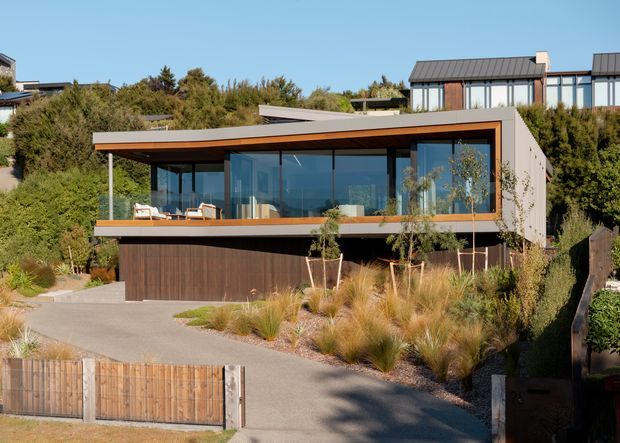 Contemporary multi-level hillside residence with a broad low-pitched roof, extensive floor-to-ceiling glazing, warm timber soffit and balcony detailing, dark vertical timber cladding, and a landscaped driveway. The home is surrounded by established vegetation and ornamental grasses, with a neighbouring modern dwelling visible behind.