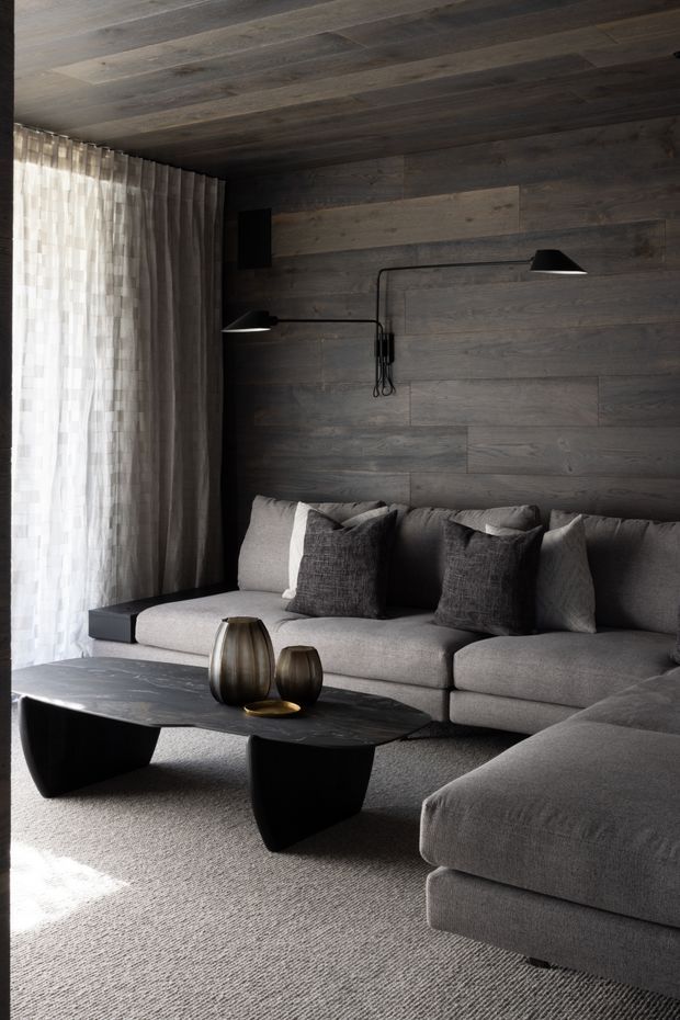 A moody, contemporary living room with dark horizontal timber wall and ceiling panelling, a large grey sectional sofa, black sculptural coffee tables, sheer floor-to-ceiling curtains, and minimalist black wall-mounted lamps. The room has a restrained monochromatic palette with warm natural timber tones and soft daylight.