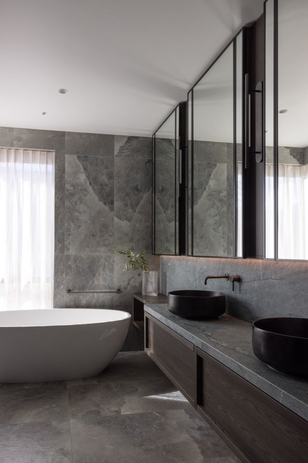 A refined contemporary bathroom featuring a freestanding white bathtub, double dark vessel basins on a long timber vanity, and a grey stone-clad wall and floor. Large mirrored cabinetry with slim black metal framing spans the vanity wall, while sheer curtains provide soft natural light. The palette is subdued grey, charcoal, black, and dark timber, creating a luxurious, calm atmosphere.