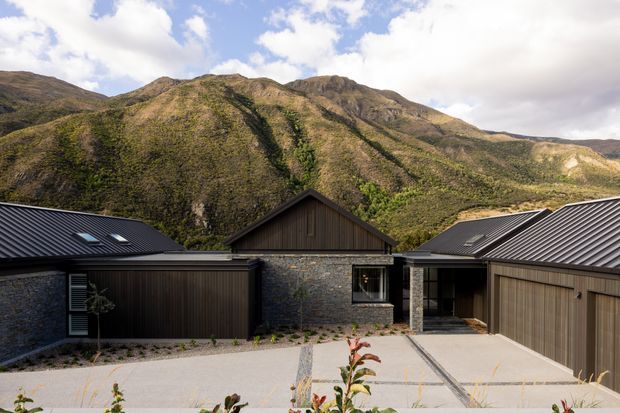 Exterior view of a contemporary dark-clad mountain residence arranged around a broad paved driveway or courtyard. The home features charcoal metal pitched roofs, dark vertical cladding, and a central section faced in stacked natural stone, set directly against a rugged green mountain landscape.