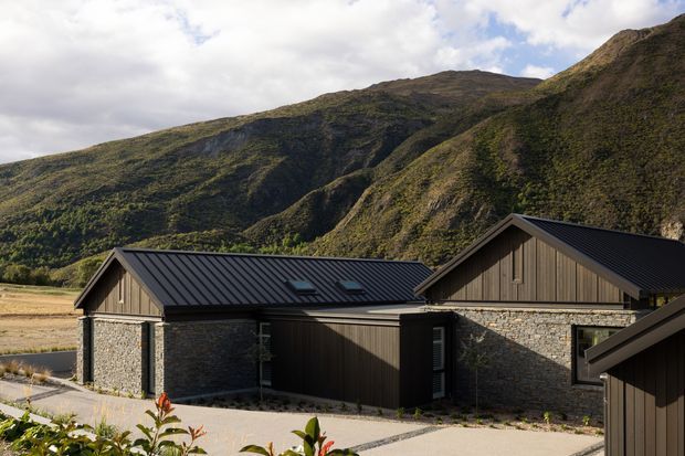 Exterior view of a low, multi-wing building set against steep mountainous terrain. The structure has dark standing-seam pitched roofs, vertical timber-clad gable sections, and substantial rough stone masonry walls, with skylights visible on the central roof. Paved paths and sparse landscaped planting surround the building.