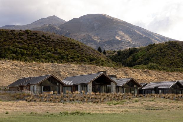 Wide exterior view of a low, single-storey mountain lodge or residential complex set against a rugged alpine mountain landscape. The buildings feature repeated pitched gable roofs with dark metal roofing, extensive glazing, and grey masonry or stone-like walls, arranged across a dry grassland setting.
