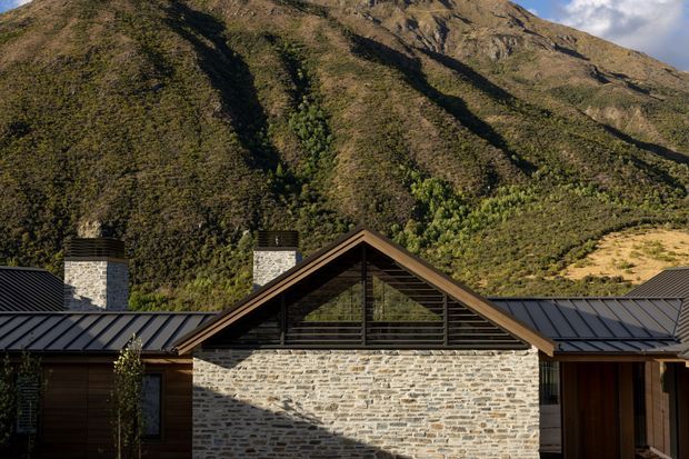 Exterior architectural photograph of a low, mountain-set building with a prominent central gabled volume. The facade combines pale rough stone masonry, dark timber framing and louvred glazing, with dark standing-seam metal roofs and two stone chimneys. Steep vegetated mountain slopes rise directly behind the building.