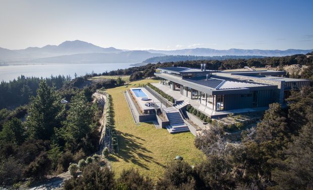 Aerial view of a large contemporary flat-roofed home set on a landscaped, elevated coastal or lakeside site. The building features extensive glazing, multiple low horizontal roof planes, terraces and stairs descending toward a long narrow swimming pool. A broad water body, wooded shoreline and mountainous landscape are visible beyond the property.