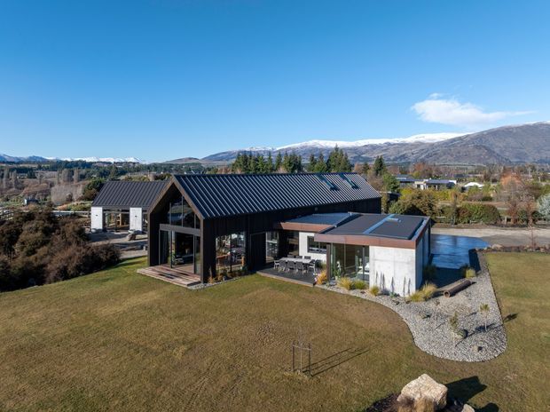 Aerial view of a contemporary single-storey home set in a spacious rural or semi-rural landscape. The house features a dark metal gabled roof, extensive floor-to-ceiling glazing, black vertical cladding, and a contrasting low flat-roofed wing with pale rendered walls. Mountain ranges and established trees form the background, while gravel paths and open lawn surround the building.