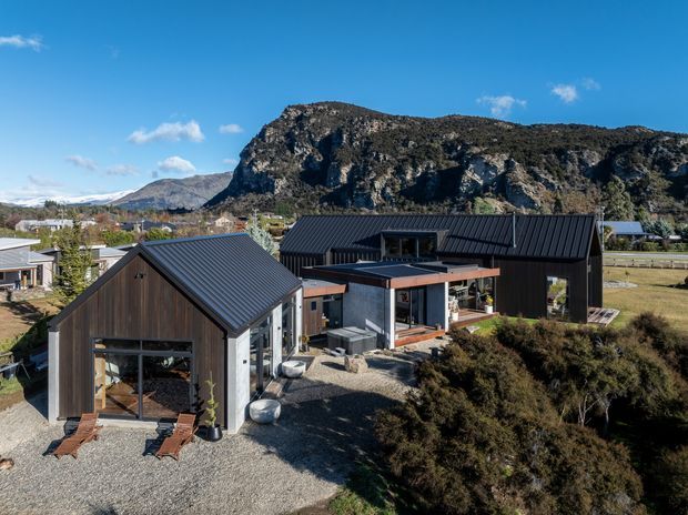A contemporary single-storey residential property viewed from above, with several connected dark-clad volumes, steep metal gable roofs, large glazed openings, and a central flat-roofed outdoor living or entry structure. The house sits in a gravelled landscaped setting with low shrub planting, open lawn, and neighbouring homes. A dramatic rocky mountain escarpment forms the backdrop under a clear blue sky.