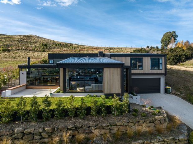 Exterior view of a contemporary single-storey hillside house with dark metal cladding, a low-pitched metal roof, extensive glazing, timber-toned vertical wall cladding, and an integrated garage. The home is set into a landscaped sloping site with stone retaining walls and clipped hedges.