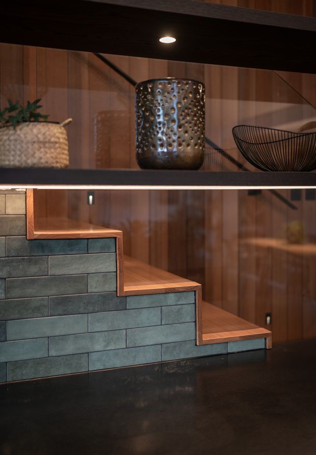 A close-up architectural interior detail showing stepped stair-like built-in joinery clad in dark blue-green rectangular tiles, with warm timber treads and shelving. The background features rich vertical timber wall panelling, dark horizontal shelves, integrated lighting, and curated decorative objects including a woven planter, metallic vessel, and wire bowl. The overall composition is warm, tactile, and refined with strong material contrast.