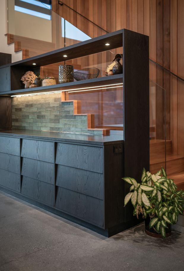 A refined open-plan interior featuring a dark timber built-in kitchen or bar joinery unit with integrated open shelving and under-shelf lighting. The cabinetry has deep blue-grey timber fronts and a dark stone benchtop, paired with a green-grey textured tile splashback. Warm timber wall panelling, a staircase, decorative objects, and a potted plant create a sophisticated, moody atmosphere.