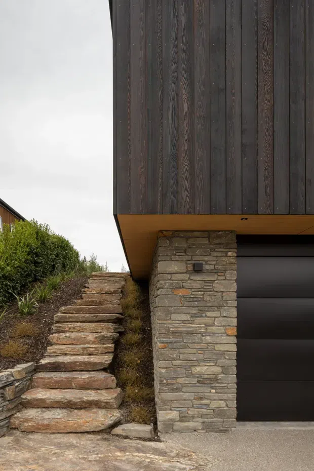 Close-up exterior view of a contemporary house entrance and garage. Dark vertical timber cladding forms the upper facade, supported by a stacked natural-stone pier. A black sectional garage door sits beneath a projecting timber-lined soffit, while rough stone steps rise through a landscaped slope alongside the building.