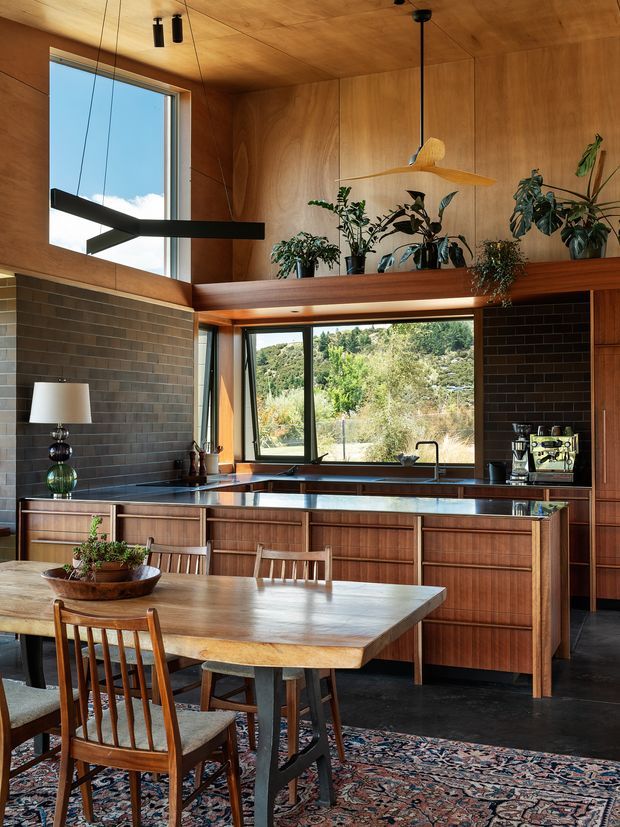 A warm, wood-lined kitchen and dining area with rich timber cabinetry, dark rectangular brick tile walls, a dark stone benchtop, and large windows overlooking greenery. The kitchen includes an island with integrated seating, sink, cooktop, coffee machine, and hanging pendant lights. A timber dining table and chairs sit on a patterned rug in the foreground, while plants and sculptural lighting add character.