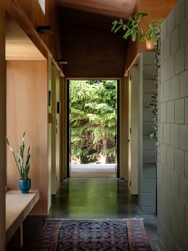 A narrow, warmly lit hallway leading to a glazed exterior door and lush greenery beyond. The corridor features extensive natural timber cabinetry and wall panelling, a dark timber ceiling, polished concrete flooring, a grey painted concrete-block wall, recessed lighting, built-in bench seating, and potted plants. A patterned runner adds colour and texture in the foreground.