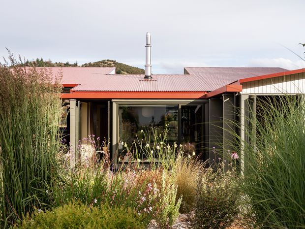Single-storey contemporary house viewed from a landscaped garden, with a low-pitched red metal roof, dark exterior walls, extensive glazing, and a prominent metal chimney flue. Tall ornamental grasses and flowering planting partially screen the facade, while a wooded hillside is visible behind the property.