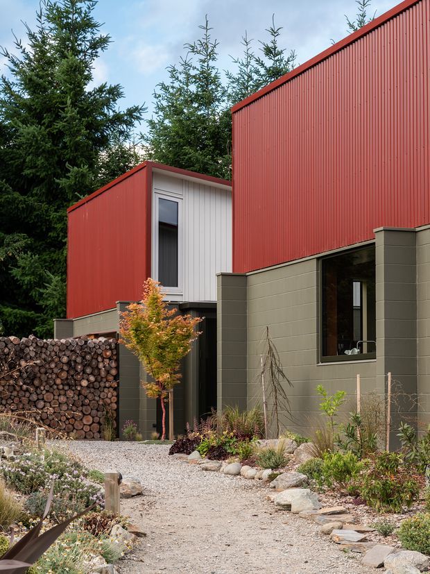 Exterior view of a contemporary multi-volume building set among mature evergreen trees. The architecture features bold red vertically ribbed metal cladding, muted olive-grey masonry walls, dark-framed glazing, and a gravel garden path with drought-tolerant landscaping and a stacked log feature wall.
