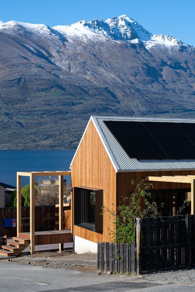 Exterior view of a contemporary timber-clad house with a steep gabled roof, dark roof-mounted solar panels, and a timber-framed entry deck. The house sits beside a blue lake, with a dramatic snow-capped mountain range visible behind it. The facade combines warm vertical timber cladding, dark window frames, and a black fence, creating a crisp alpine setting.