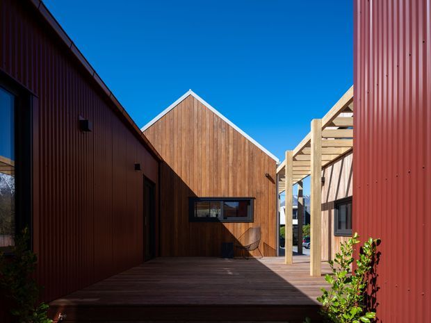 A sunny architectural exterior courtyard or passage between timber-clad buildings. The composition features dark red corrugated metal wall cladding on both sides, a warm vertical timber gable wall at the end, a timber deck, and a light timber pergola-like overhead structure. Strong sunlight and deep shadows create a crisp, contemporary residential setting.