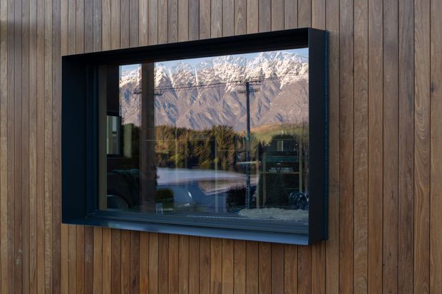 Close-up exterior view of a dark-framed rectangular window set into vertical timber wall cladding. The reflective glazing shows a dramatic snow-capped mountain range and hints of an interior living space, while the timber facade has a warm, weathered natural finish.