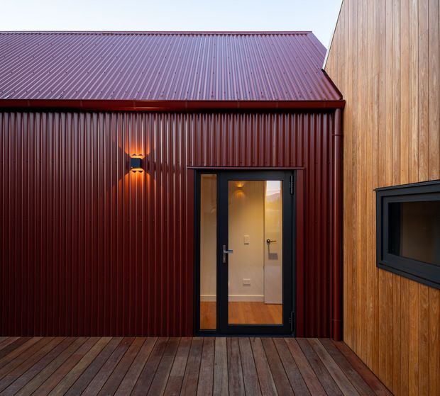 A compact modern courtyard or deck-facing exterior with a deep burgundy corrugated metal-clad wall and pitched metal roof, contrasted with warm vertical timber cladding on an adjoining wall. A black-framed glazed entry door opens into a warmly lit interior, while a black-framed horizontal window is visible in the timber-clad wall. Timber decking spans the foreground and wall-mounted lighting adds a warm glow.