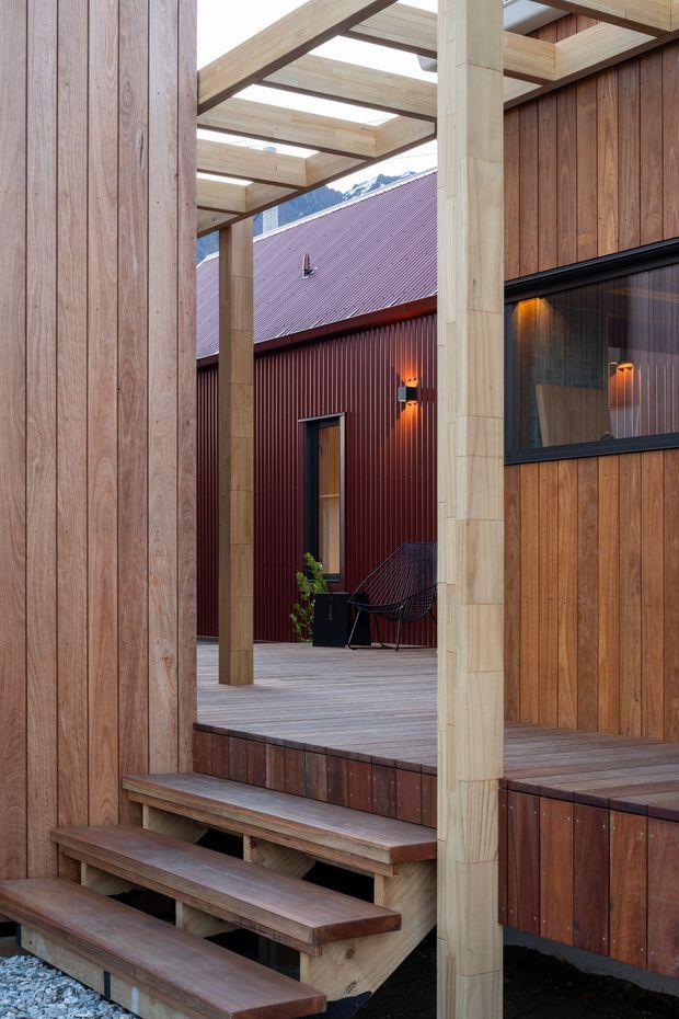 A timber-clad courtyard or outdoor transition space between buildings, with broad wooden steps leading to a raised deck. Pale exposed timber posts and beams form an open pergola overhead, while darker vertical timber cladding and a deep red corrugated metal building surround the space. Warm wall lights, a narrow window, outdoor seating, and a mountainous backdrop are visible.