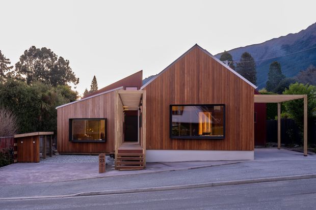 A compact contemporary single-storey house viewed from the street, with a strongly expressed steep gabled volume clad in vertical warm timber boards. The facade includes dark-framed windows, a recessed entry with timber steps, a low adjoining wing, and a simple covered carport or outdoor canopy to the right. Mountainous terrain and mature vegetation form the background.