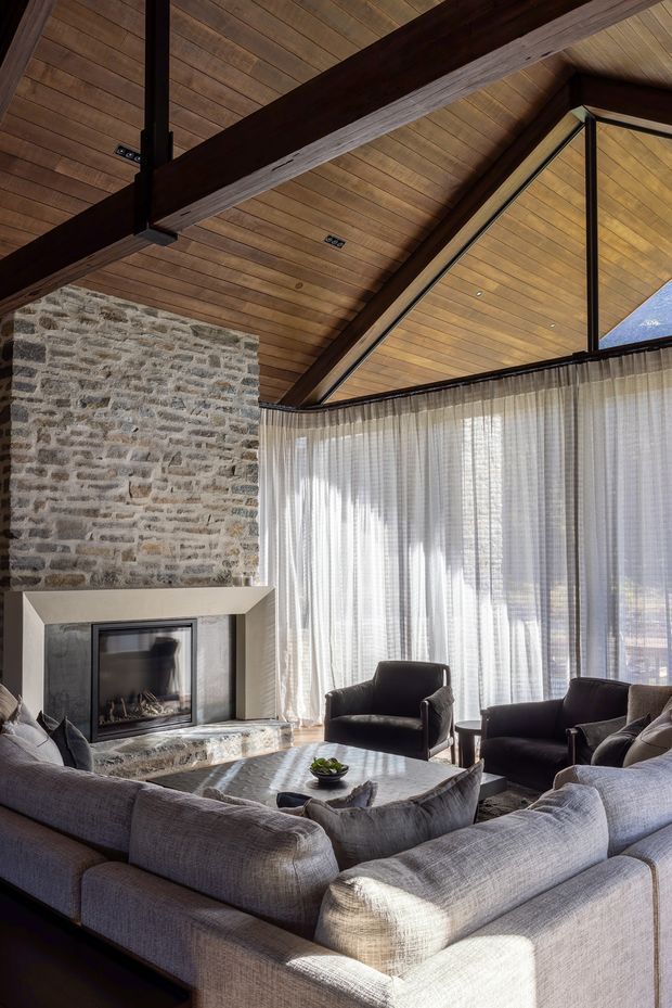 A refined living room with a dramatic vaulted timber ceiling, exposed dark timber beams, a textured natural-stone fireplace wall, and expansive glazed openings dressed with sheer curtains. A large light-grey sectional surrounds a substantial irregular stone coffee table, with dark lounge chairs arranged near the fireplace. The space has strong natural light and a warm, lodge-like yet contemporary character.