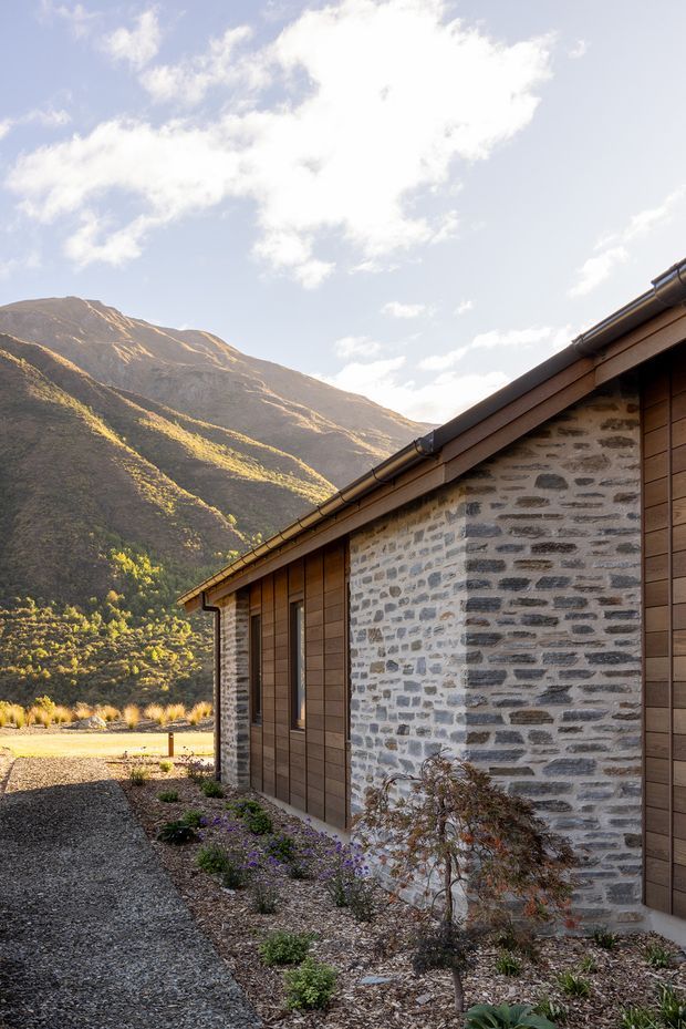 Exterior view of a low-slung mountain building with grey natural stone masonry walls, warm brown vertical timber cladding, dark metal gutters and a shallow pitched roof. A gravel path and sparse landscaped planting run alongside the facade, with a dramatic alpine mountain backdrop in warm late-day light.