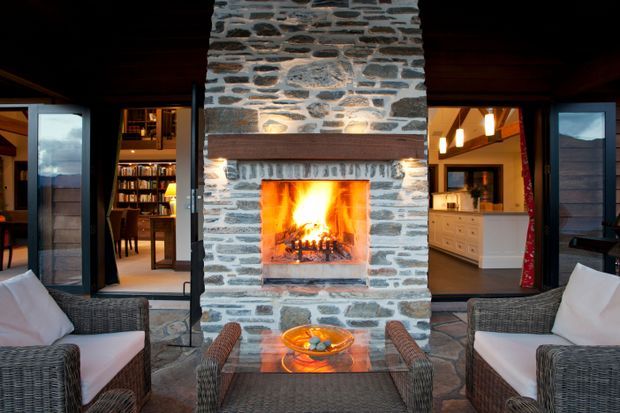 Interior view of a warm, open-plan living space centred on a large exposed natural-stone fireplace with a lit fire and timber mantel. Wicker armchairs and a glass coffee table occupy the foreground, while open doorways reveal a library or dining area on the left and a kitchen with pale cabinetry on the right. Dark timber ceilings and framing create a cosy lodge-like atmosphere.