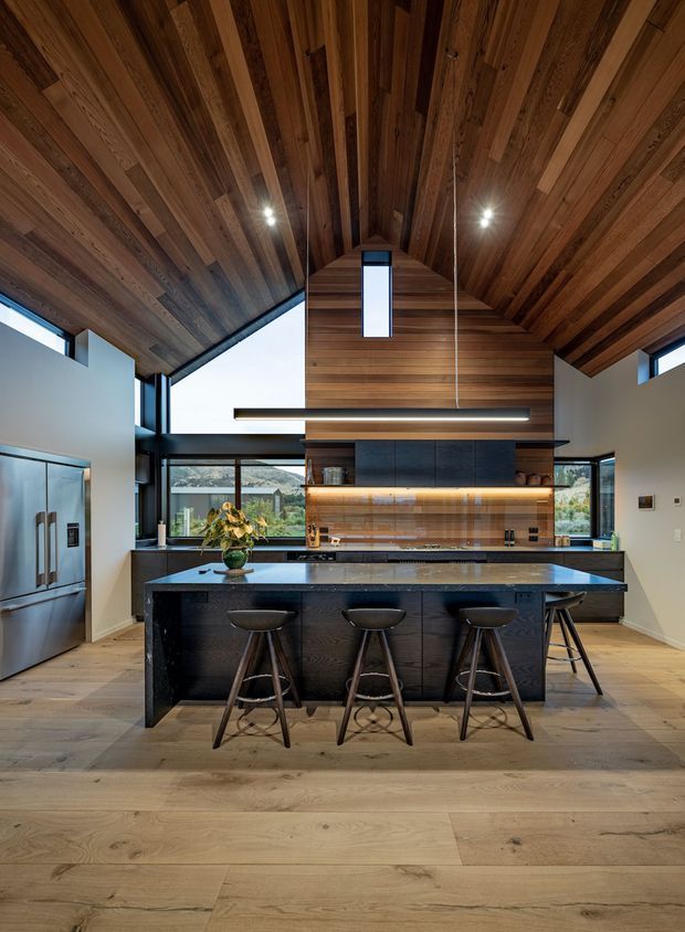 A spacious contemporary kitchen with a large dark island and seating for four, integrated stainless-steel appliances, extensive timber wall and vaulted ceiling panelling, dark stone benchtops, and warm timber flooring. Large windows and clerestory glazing provide natural light, while linear pendant lighting illuminates the island. The kitchen opens toward a glazed living or dining area beyond.