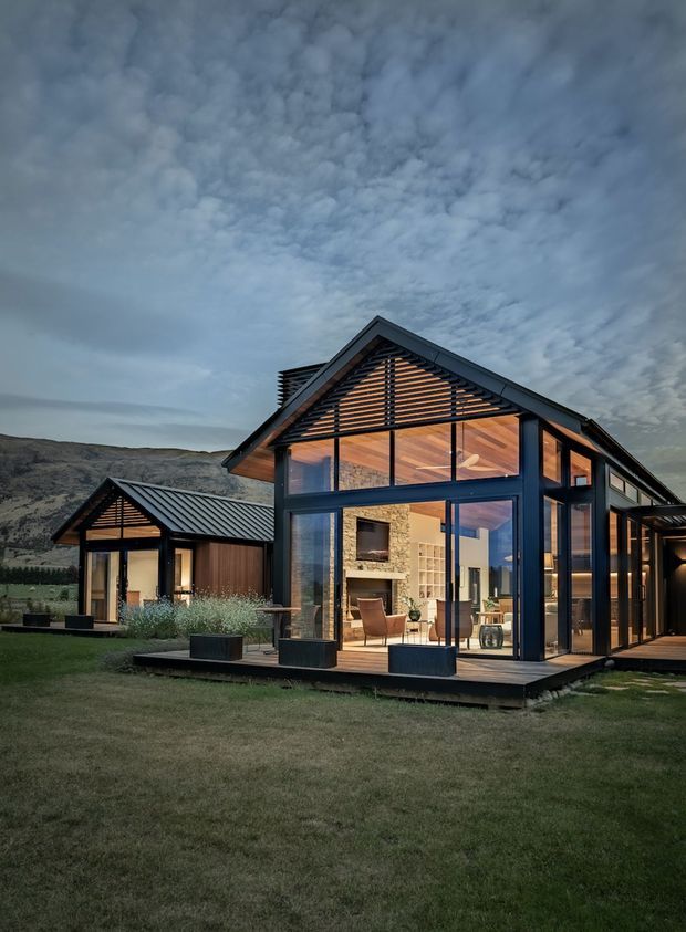 Exterior dusk view of a modern single-storey pavilion-style house set against a rugged hillside. The main gabled volume has extensive floor-to-ceiling glazing, dark metal framing, a projecting timber-lined roof, and warm interior lighting. A smaller matching gabled structure sits to the left, connected by a deck and landscaped lawn. The transparent facade reveals an open living area with a stone feature wall and timber ceiling.