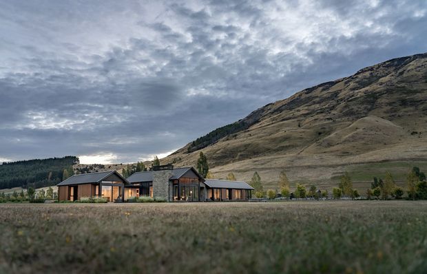 A low, modern rural residence set in an open grassland landscape at the foot of a large dry, rolling mountain. The home has a horizontally composed, single-storey form with dark metal pitched roofs, extensive glazing, dark vertical cladding, and a prominent central stone-clad volume. The overcast sky and muted vegetation create a calm, natural atmosphere.