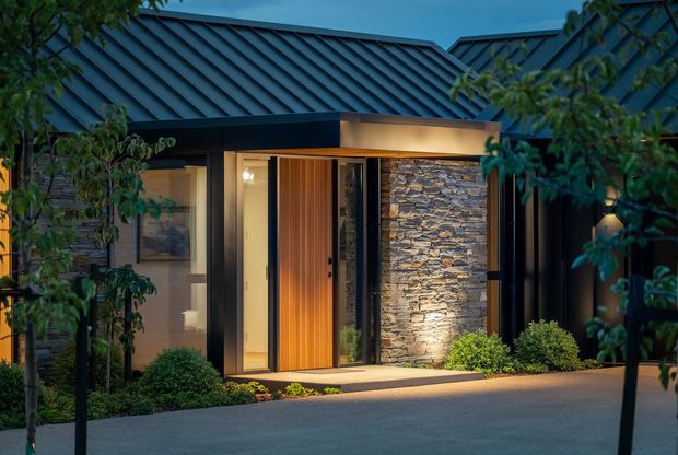 A contemporary single-storey home entrance viewed from outside at dusk. The facade combines dark metal cladding and a standing-seam pitched roof with natural stacked-stone walling, warm timber entry detailing, full-height glazing, and a sheltered porch. Soft exterior lighting illuminates the entry and low landscaped planting surrounds the paved approach.