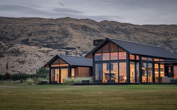 A contemporary single-storey mountain residence viewed from across a broad lawn. The building consists of connected gabled volumes with dark vertical cladding, charcoal standing-seam metal roofs, extensive floor-to-ceiling glazing, and warm interior lighting. A rugged, sparsely vegetated mountain landscape forms the backdrop.
