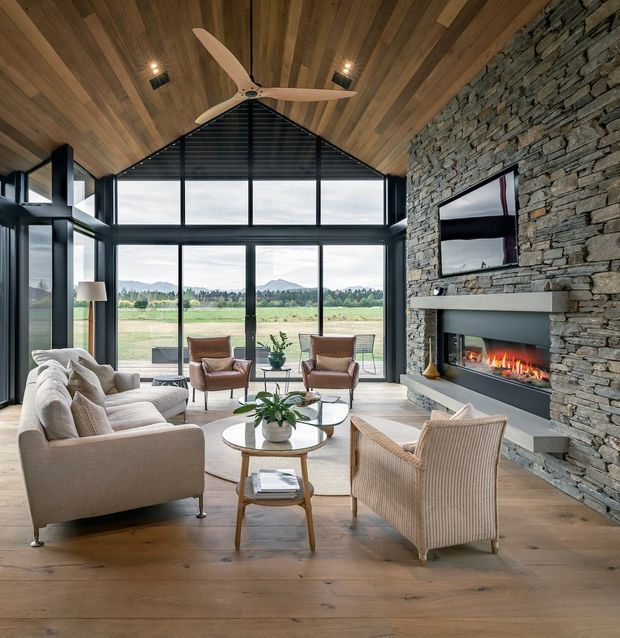 A spacious, light-filled living room with a dramatic vaulted timber-lined ceiling, exposed gable form, black-framed floor-to-ceiling glazing, and a rugged stacked-stone fireplace wall. Neutral upholstered sofas and armchairs surround round coffee tables, while a large ceiling fan hangs centrally. The glazing opens views toward fields and distant hills, creating a warm, refined indoor-outdoor atmosphere.