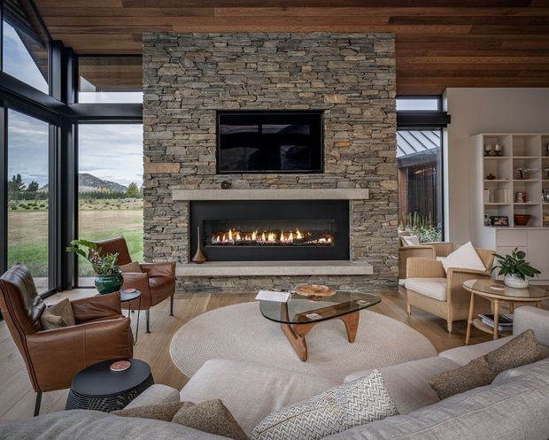 A spacious, refined living room centred on a tall stacked-stone fireplace wall with a built-in linear fire and television niche. Floor-to-ceiling black-framed glazing opens to a rural landscape, while a timber-lined ceiling, built-in shelving, leather lounge chairs, a large sectional sofa, glass coffee table and layered neutral furnishings create a warm contemporary lodge-like interior.