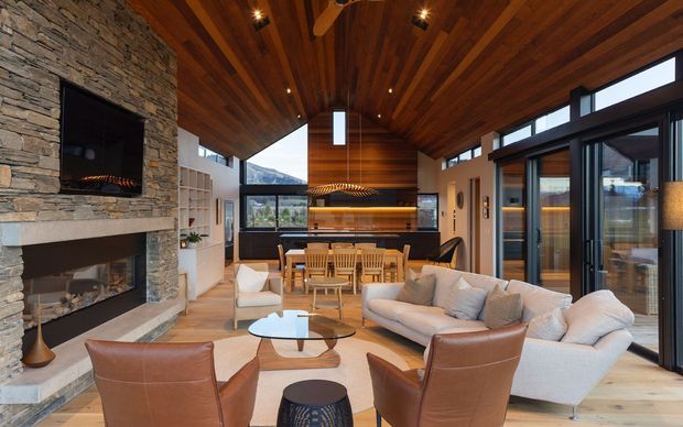 A spacious, open-plan living room with a dramatic pitched timber-lined ceiling, extensive dark-framed glazing, a large stacked-stone fireplace wall, and an integrated kitchen and dining area. The room is furnished with a pale sectional sofa, leather lounge chairs, a glass coffee table, and timber dining furniture. The glazing opens toward a wooded or mountainous landscape, while warm timber and stone finishes create a refined lodge-like atmosphere.