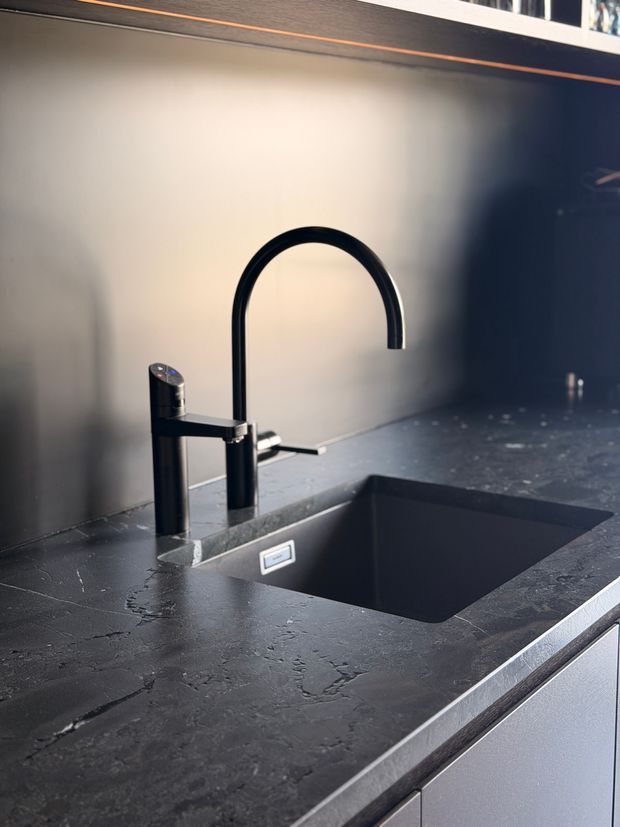 Close-up view of a contemporary kitchen sink area, featuring a matte-black undermount sink, tall black gooseneck mixer tap, dark veined stone benchtop, and dark cabinetry with a minimal integrated splashback.