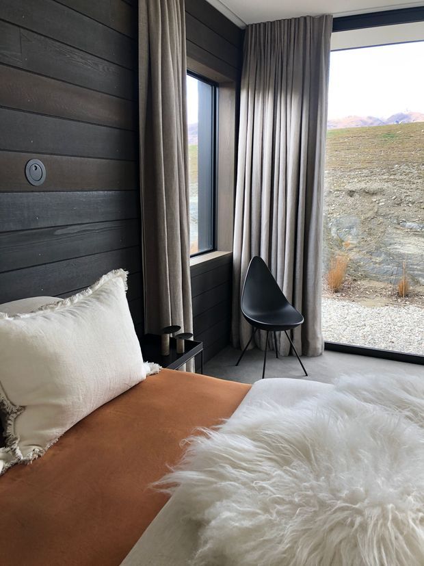 A compact, contemporary bedroom with dark horizontal timber wall panelling, floor-to-ceiling glazing, and full-height taupe curtains. The room contains a low bed with a warm tan cover, cream textured cushions, a white sheepskin throw, a small dark bedside table, and a black sculptural chair. A rugged mountainous landscape is visible outside.
