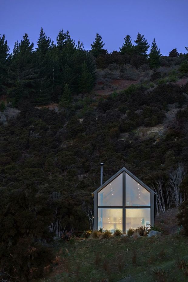 A small, narrow modern glass house sits at the base of a densely vegetated hillside, photographed at dusk. The compact dwelling has a steep gabled roof, dark structural framing, extensive floor-to-ceiling glazing, and warm interior lighting visible through the facade. Native-looking shrubs and trees surround the building on a sloping site.