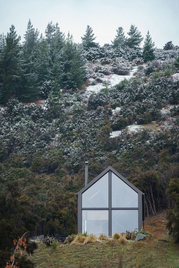 A compact contemporary cabin or studio sits at the base of a steep, densely vegetated hillside dusted with snow. The front facade is dominated by tall, geometric floor-to-roof glazing divided by dark structural frames, with a simple pitched gable roof and a visible metal chimney. Native grasses and low planting surround the building, creating a secluded alpine or bush setting.