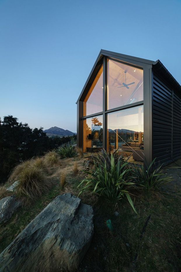 Exterior architectural photograph of a compact contemporary building set into a rugged landscaped site. The dark-clad structure has a steep gabled glazed end wall with expansive floor-to-roof glazing, a visible ceiling fan and warm interior lighting. The side elevation features horizontal dark slatted cladding and a dark metal roof edge. Native grasses, spiky planting and exposed rocks surround the building, with a mountainous landscape visible beyond.