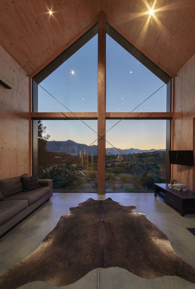 A dramatic double-height living room with a steeply pitched timber-lined ceiling and exposed central timber post-and-beam structure. Floor-to-ceiling triangular and rectangular glazing frames a mountain landscape at dusk. The room has polished concrete flooring, a large natural hide rug, a low dark timber media console, and a sectional sofa. Recessed spotlights and the expansive glazing create a warm, refined architectural interior.